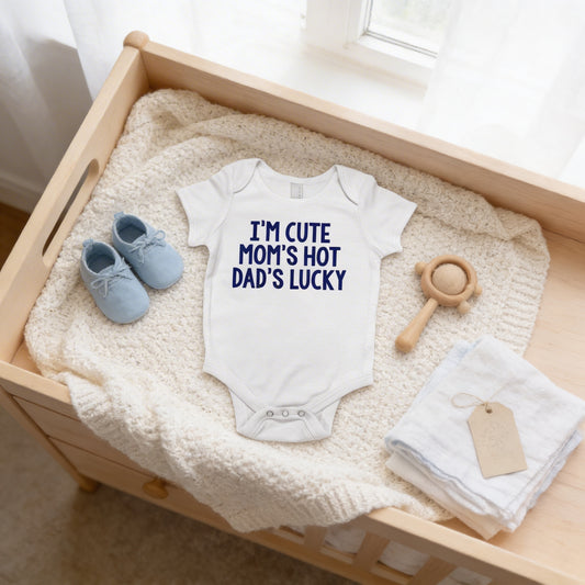 A baby's onesie with the text "I'M CUTE MOM'S HOT DAD'S LUCKY" is displayed on a wooden dresser, accompanied by a pair of blue baby shoes, a wooden rattle, and a white towel.