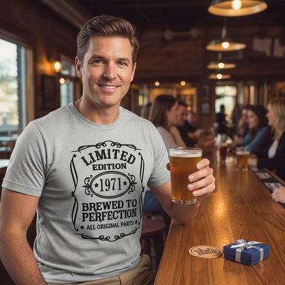 A man wearing a gray t-shirt with a design that says "Limited Edition 1971 - Brewed to Perfection All Original Parts" is sitting at a bar, holding a glass of beer.