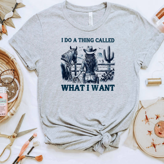 a grey t-shirt with a graphic design depicting a woman in a cowboy hat standing next to a horse, with the text "I DO A THING CALLED WHAT I WANT" printed on it.
