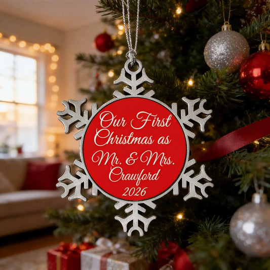 A red and silver snowflake ornament hangs from a Christmas tree, with a message that reads "Our First Christmas as Mr. & Mrs. Crawford 2026".
