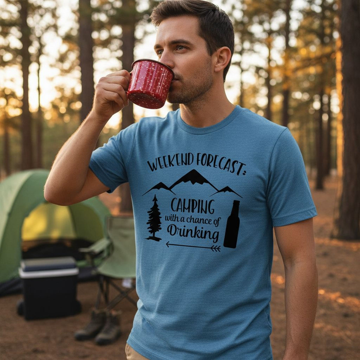 A man wearing a blue t-shirt with a graphic design stands in a forest, holding a red mug and drinking from it.