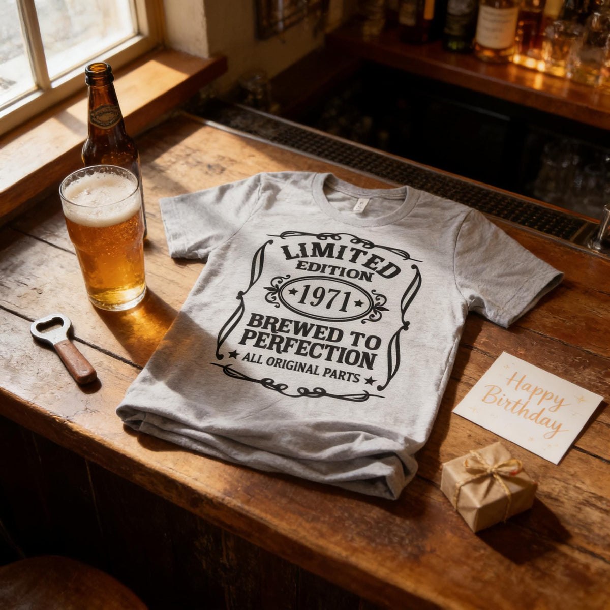 a grey t-shirt with a black and white design, a bottle of beer, a glass, a bottle opener, and a birthday card on a wooden table.