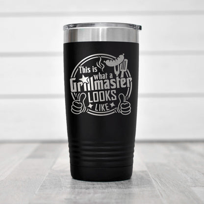 A black insulated tumbler with a silver lid, featuring a humorous text that says "This is what a Grillmaster looks like" along with a thumbs up symbol.