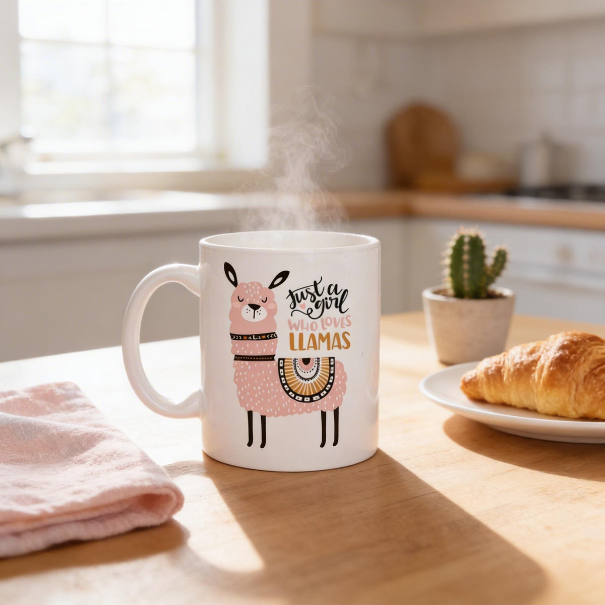 A mug with a cartoon image of a llama and the text "Just a girl who loves llamas" sits on a kitchen counter, with a plate of croissants and a potted cactus in the background.