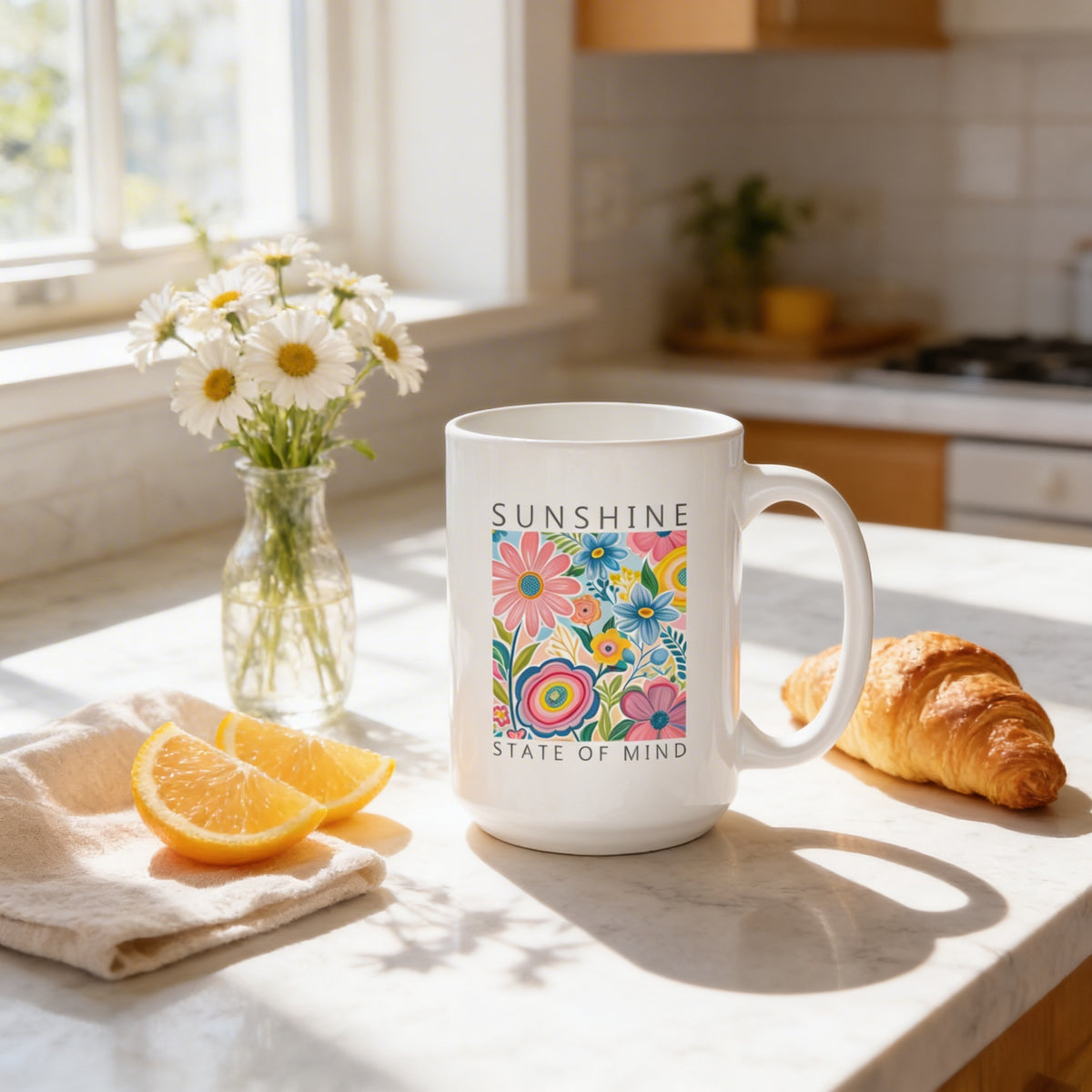 A white mug with a colorful floral design sits on a kitchen counter, accompanied by a vase of white daisies, a slice of orange, and a croissant.