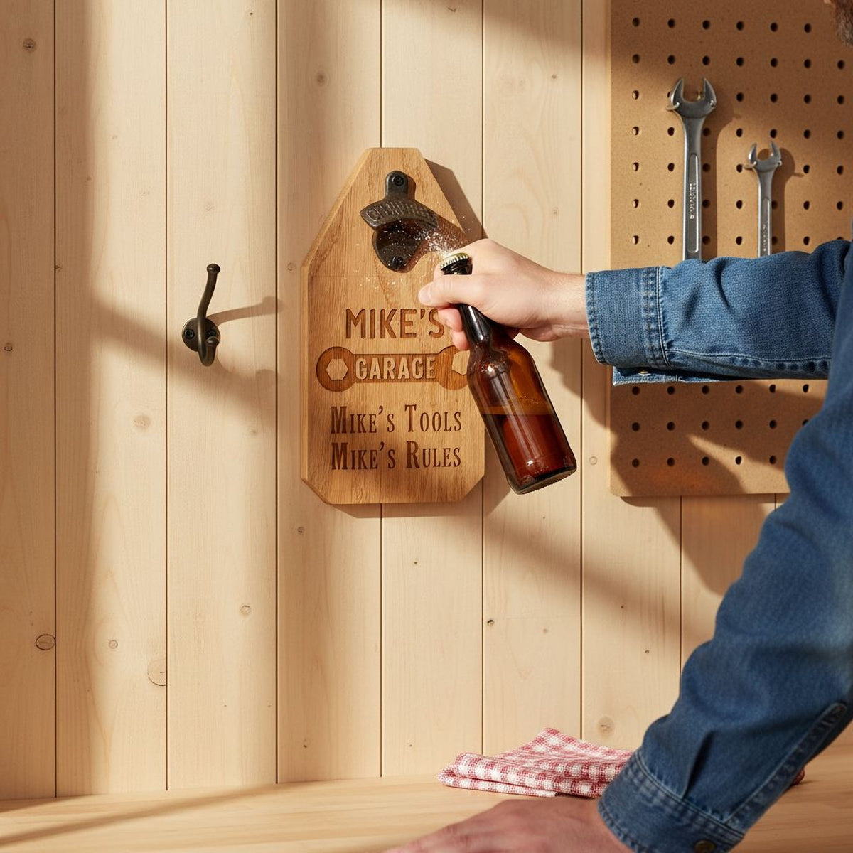 A person is holding a bottle of beer and using a wooden beer bottle opener with the inscription "Mike's Garage" on it.
