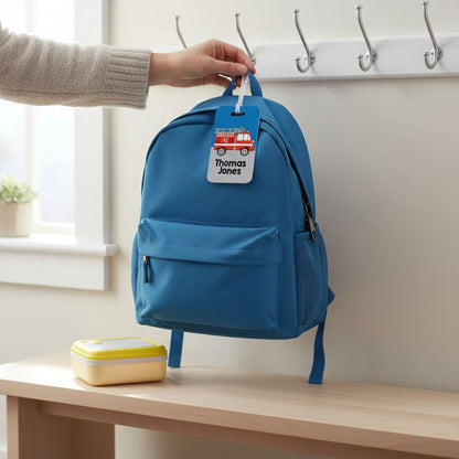 A person's hand is holding a blue backpack with a personalized name tag attached to it, which says "Thomas Jones". The backpack is placed on a wooden bench, and there is a yellow and white lunch box next to it.