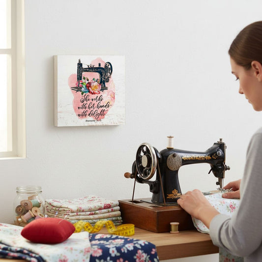 A woman is working on a sewing machine in a room with a floral print on the wall and a stack of fabric nearby.