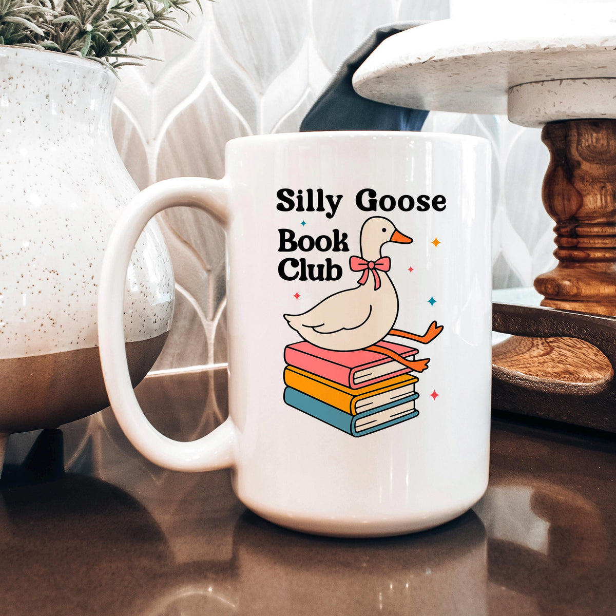 A white mug with a graphic design featuring a cartoon goose sitting on a stack of books, with the text "Silly Goose Book Club" printed on it.