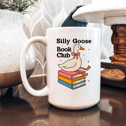 A white mug with a graphic design featuring a cartoon goose sitting on a stack of books, with the text "Silly Goose Book Club" printed on it.