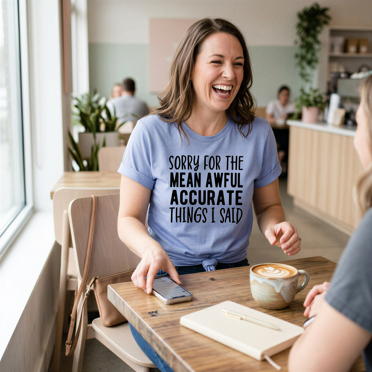 A woman wearing a blue t-shirt with a humorous text is laughing and smiling while sitting at a table in a cafe.
