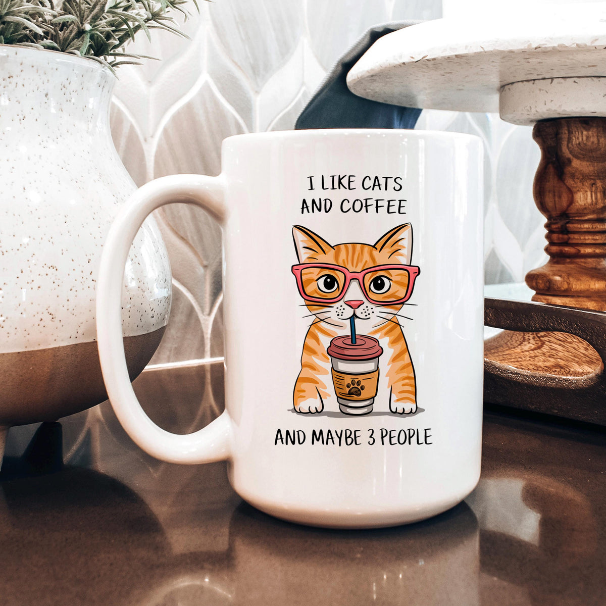 A white coffee mug with an illustration of a cat wearing glasses and holding a coffee cup, accompanied by the text "I LIKE CATS AND COFFEE AND MAYBE 3 PEOPLE".