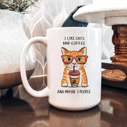 A white coffee mug with an illustration of a cat wearing glasses and holding a coffee cup, accompanied by the text "I LIKE CATS AND COFFEE AND MAYBE 3 PEOPLE".