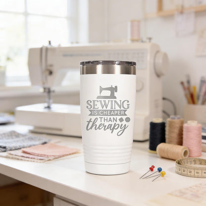 A white tumbler with a printed message on it, placed on a sewing workbench with sewing supplies and a sewing machine in the background.