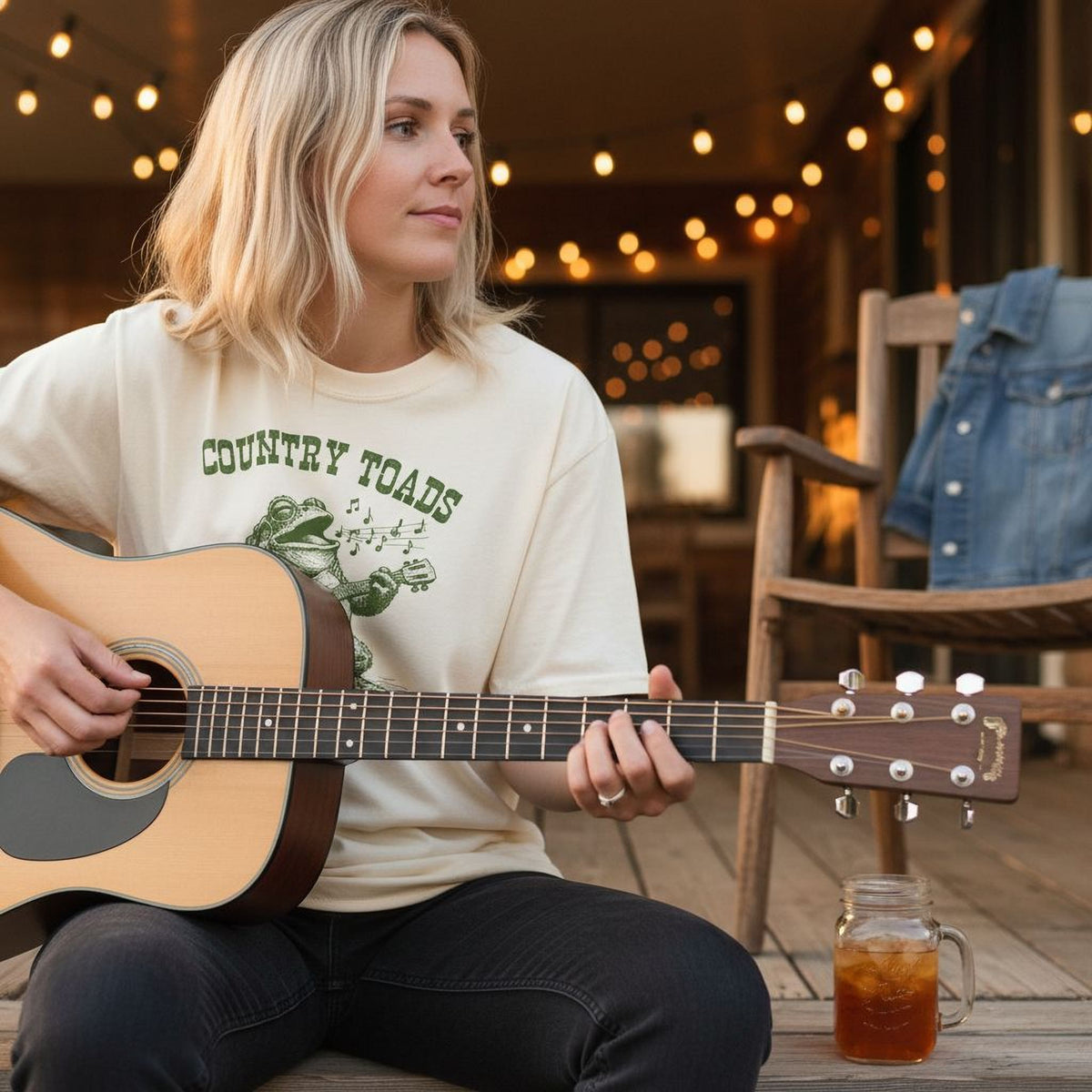 A woman is sitting on a porch, playing an acoustic guitar and holding a glass of iced tea.