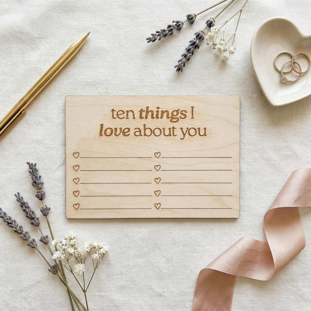 a wooden card with a list of "ten things I love about you" written on it, surrounded by various objects such as a pen, a heart-shaped dish with rings, and some flowers.