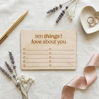 a wooden card with a list of "ten things I love about you" written on it, surrounded by various objects such as a pen, a heart-shaped dish with rings, and some flowers.