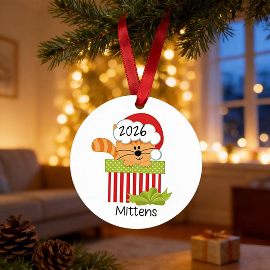 A Christmas ornament with a cat wearing a Santa hat and the number "2026" on it, hanging from a tree branch.