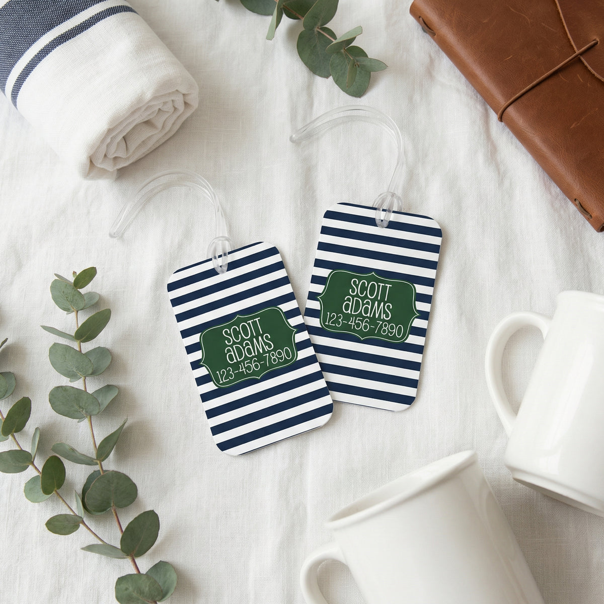 two striped luggage tags with names and phone numbers, along with some other items such as a rolled-up towel, a brown leather bag, and a white mug.