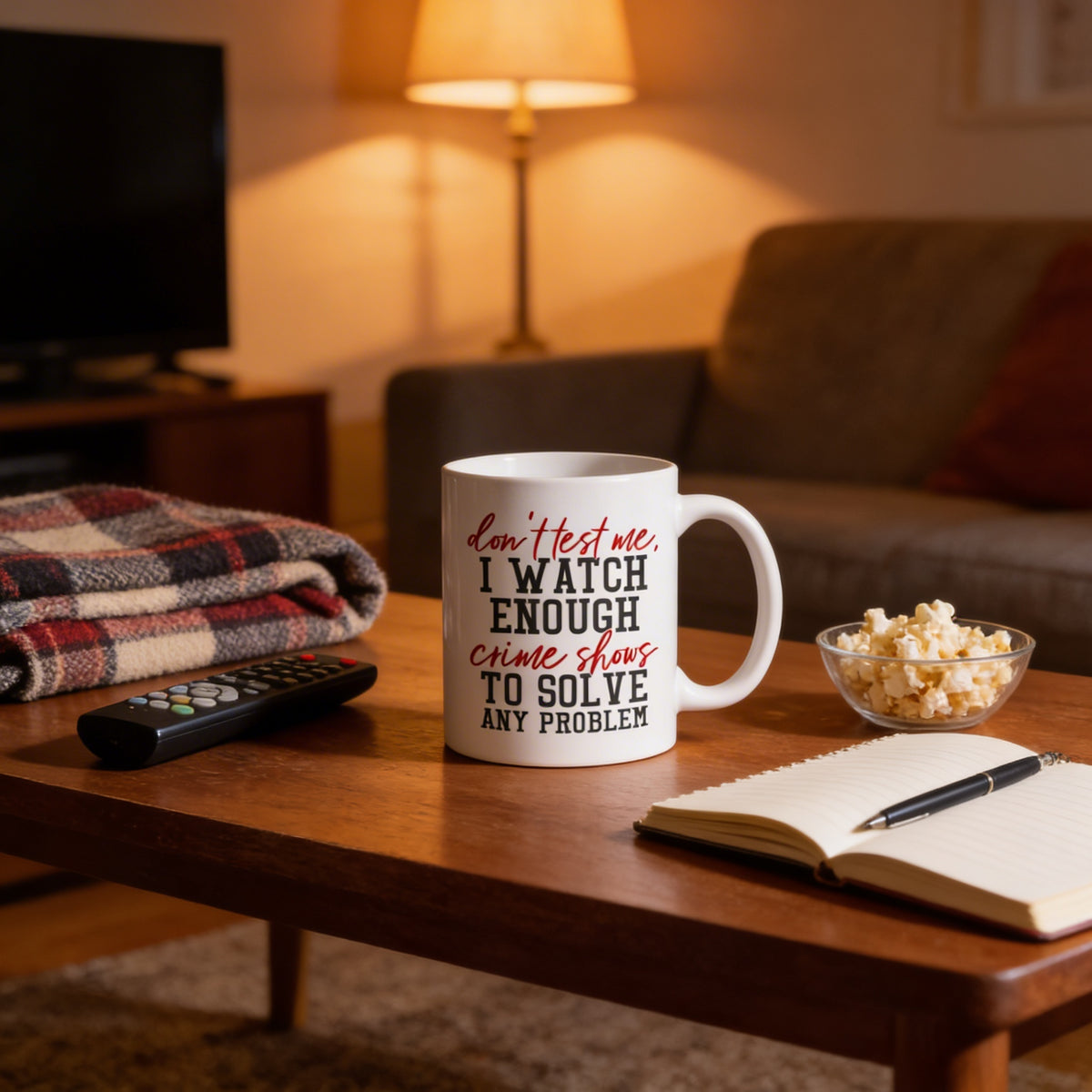 A mug with the text "don't test me, I watch enough crime shows to solve any problem" sits on a wooden coffee table, accompanied by a bowl of popcorn, a remote control, and an open notebook.