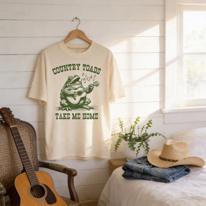 a white t-shirt with a graphic of a frog playing a guitar and the text "Country Toads Take Me Home" hanging on a wooden hanger. Next to the t-shirt, there is a guitar, a hat, and some clothing items, including a pair of jeans and a folded denim jacket.