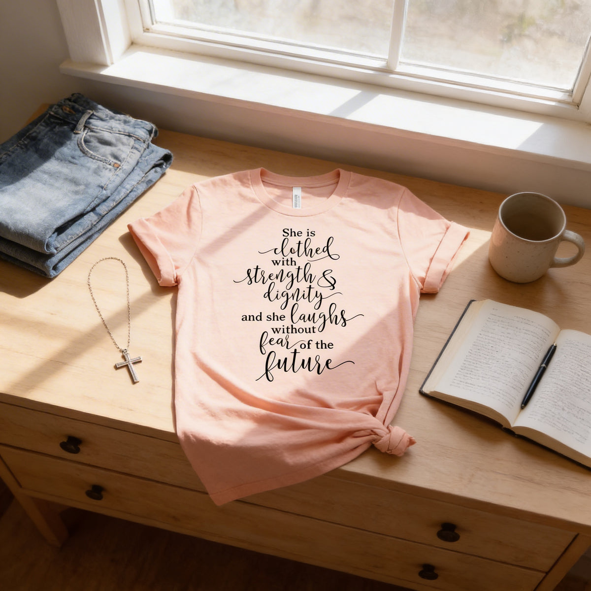 A pink t-shirt with a Bible verse printed on it, along with a cross necklace, a book, and a mug, all placed on a wooden surface near a window.