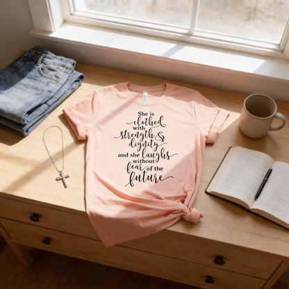 A pink t-shirt with a Bible verse printed on it, along with a cross necklace, a book, and a mug, all placed on a wooden surface near a window.