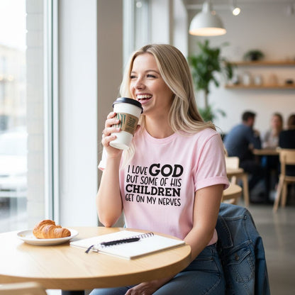A woman wearing a pink t-shirt with text is sitting at a table in a cafe, smiling and holding a coffee cup.