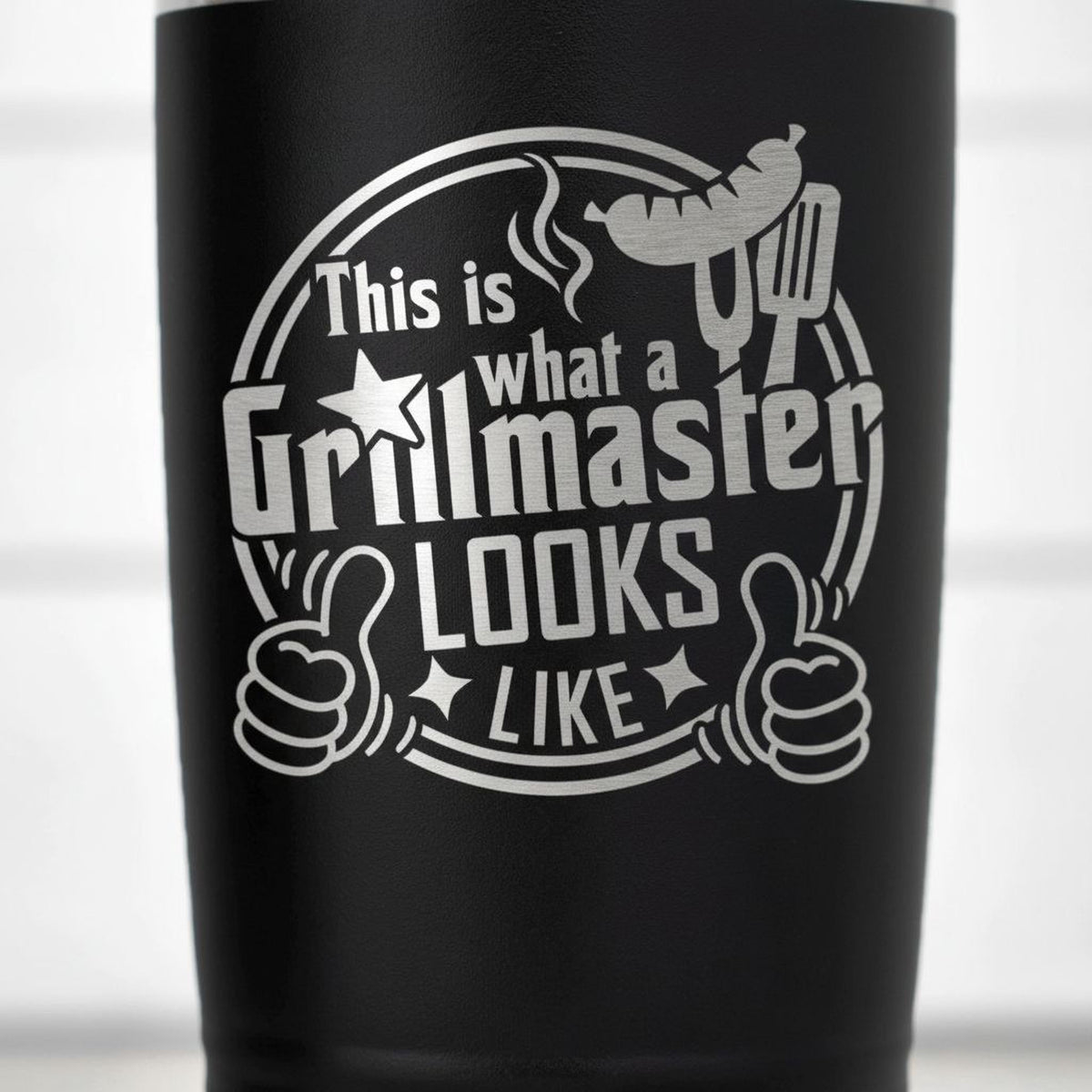 a black tumbler with a white graphic design on it. The design includes a grill, a thumbs up symbol, and the text "This is what a Grillmaster looks like like".