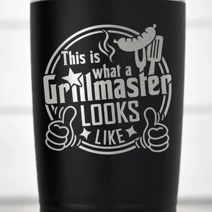 a black tumbler with a white graphic design on it. The design includes a grill, a thumbs up symbol, and the text "This is what a Grillmaster looks like like".