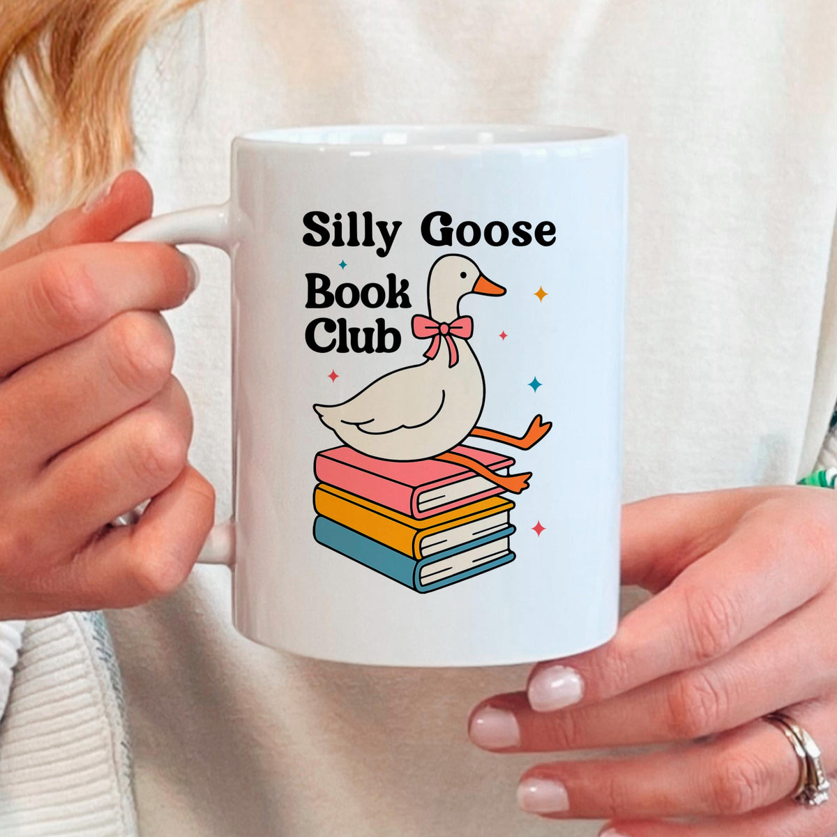 A person is holding a white mug with a graphic design featuring a cartoon goose sitting on a stack of colorful books.