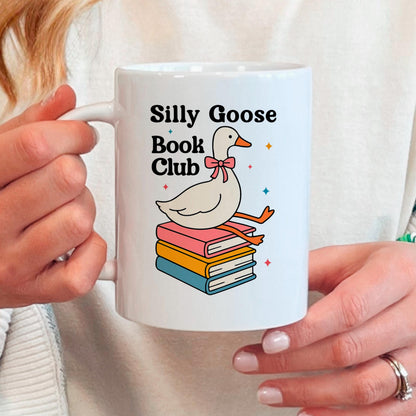 A person is holding a white mug with a graphic design featuring a cartoon goose sitting on a stack of colorful books.