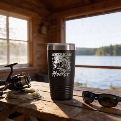 A black tumbler with a white design and text, placed on a wooden table next to a fishing rod, sunglasses, and a scenic view of a lake through a window.