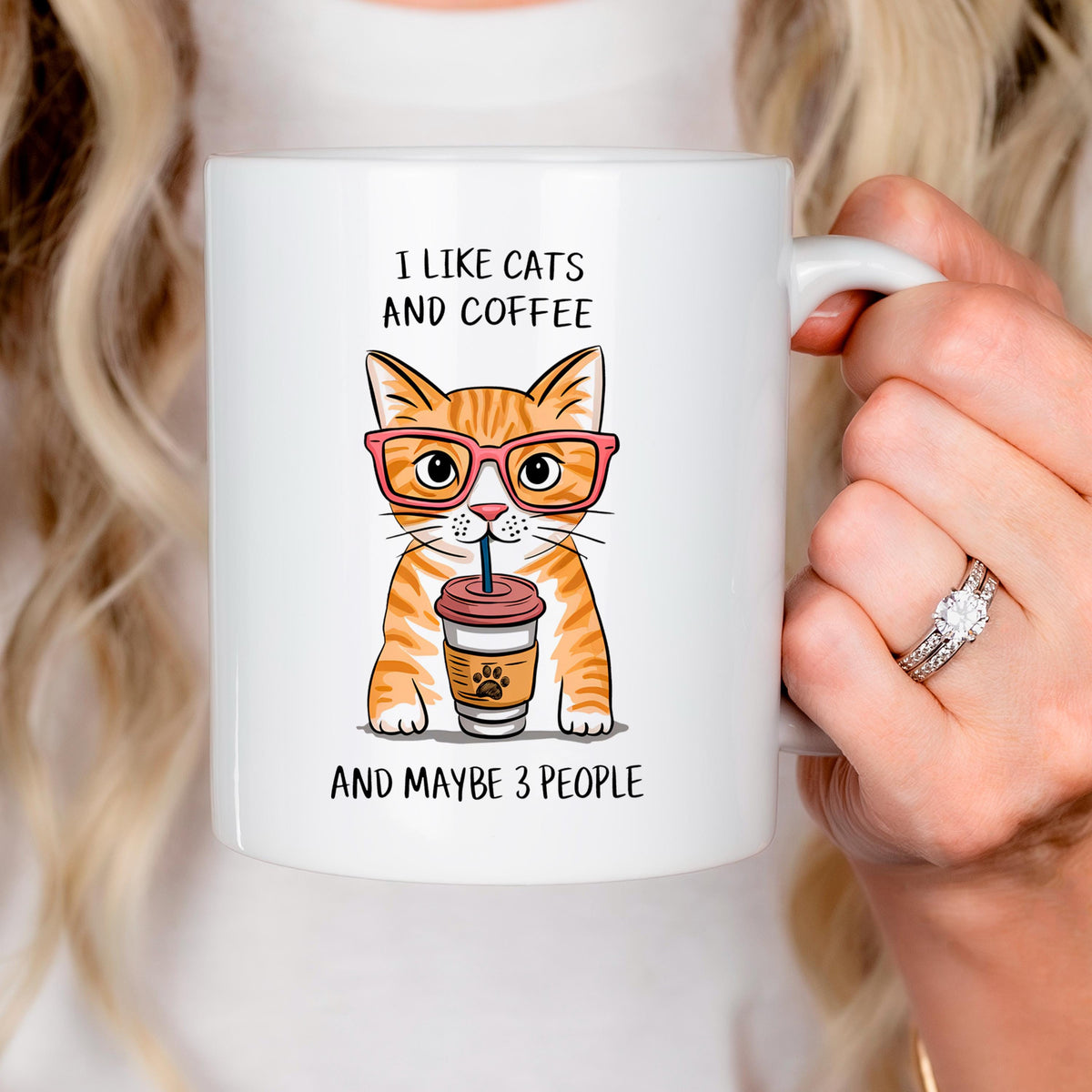 A person is holding a white mug with an illustration of a cat wearing glasses and holding a coffee cup.
