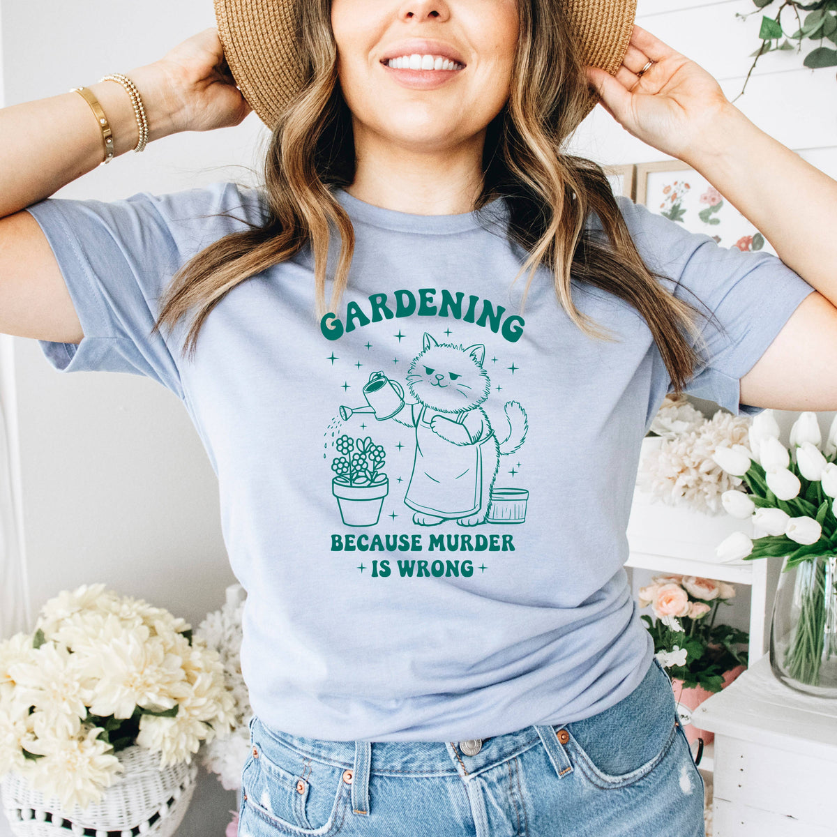 A woman wearing a gray t-shirt with a graphic design of a cat watering plants and the text "Gardening because murder is wrong".
