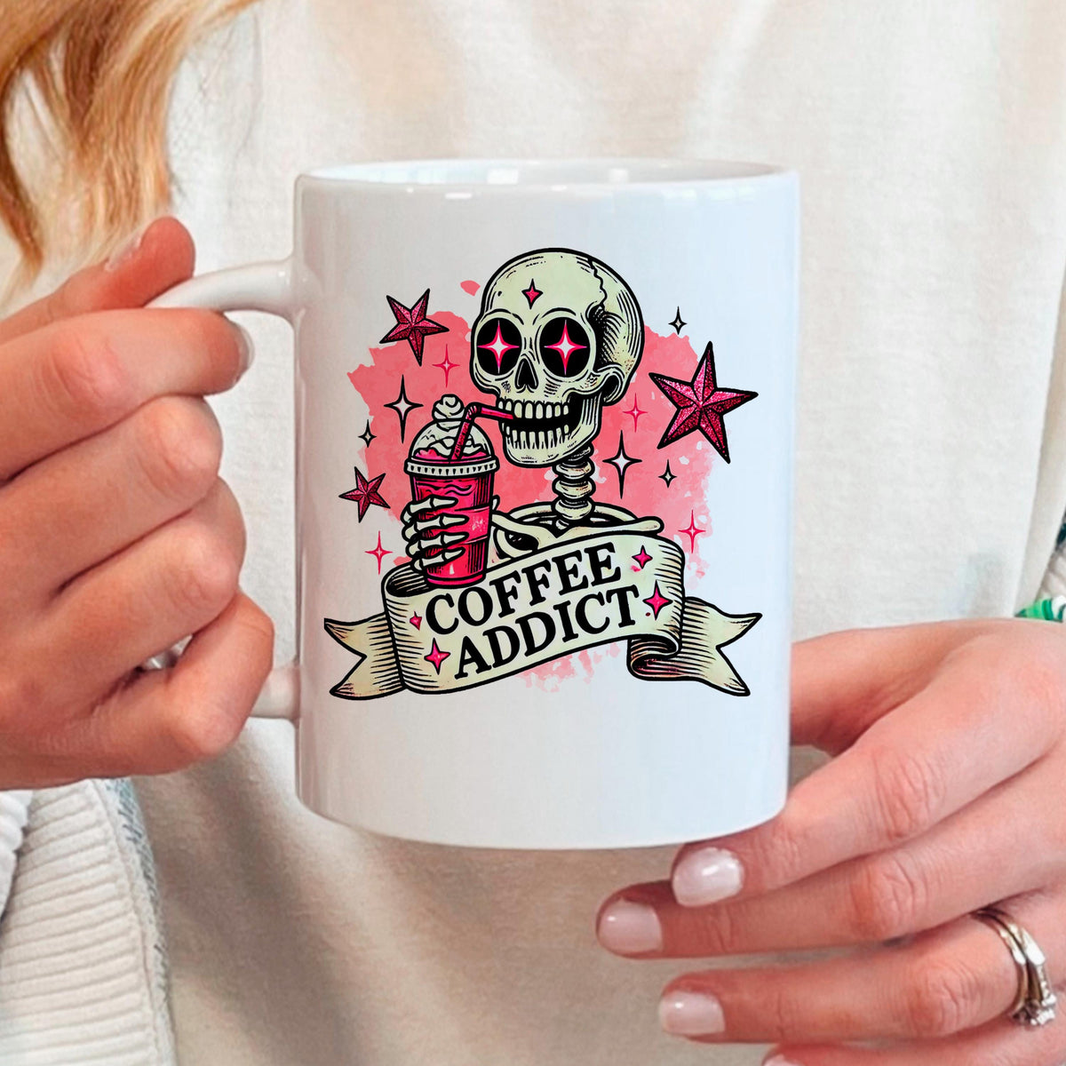 A person is holding a white mug with a graphic design featuring a skeleton holding a coffee drink and surrounded by stars and a banner that says "COFFEE ADDICT".