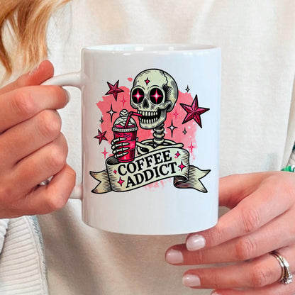 A person is holding a white mug with a graphic design featuring a skeleton holding a coffee drink and surrounded by stars and a banner that says "COFFEE ADDICT".
