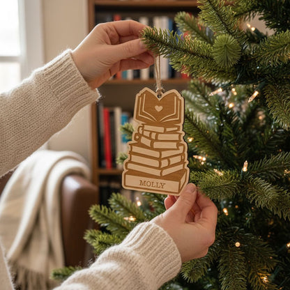 A person's hands holding a wooden ornament with a stack of books on top, hanging from a Christmas tree.