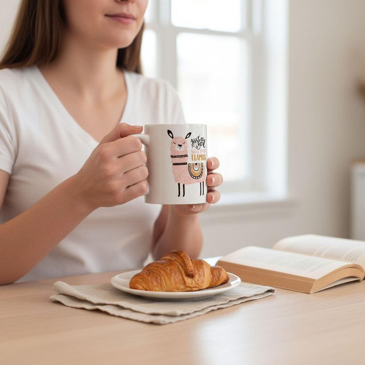 A woman is sitting at a table, holding a mug with a cartoon character on it, and enjoying a croissant and a book.