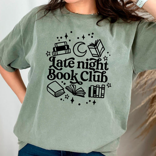 May include: A green t-shirt with a black graphic design. The design features a crescent moon, stars, and several books. The text on the shirt reads 'Late night Book Club'.