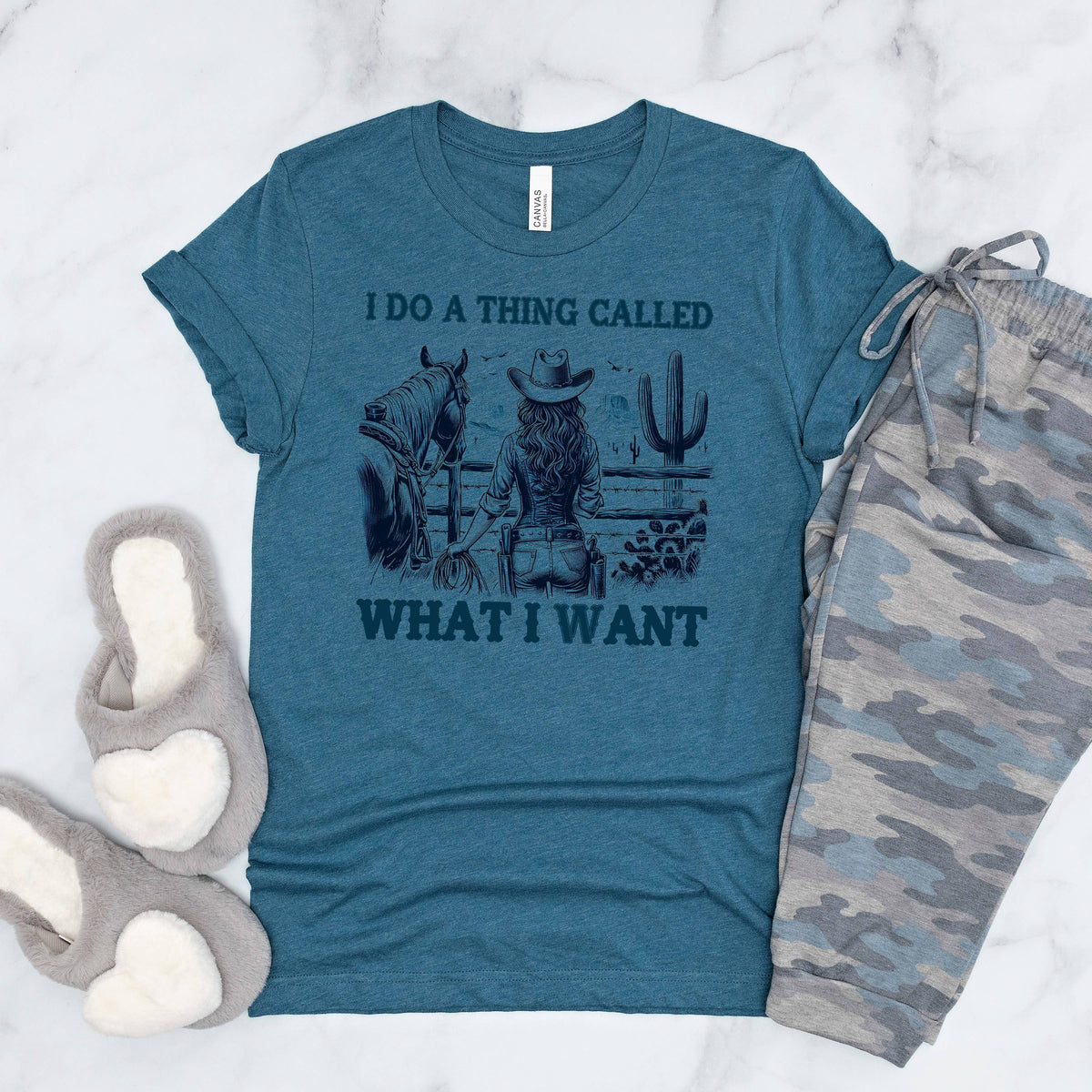 A teal t-shirt with a graphic design, a pair of camouflage pants, and a pair of gray slippers are laid out on a marble surface.