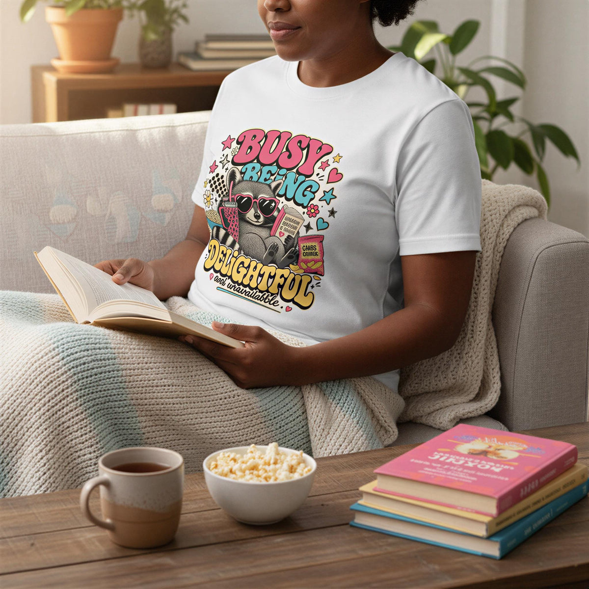 A person is sitting on a couch, reading a book and enjoying snacks, including popcorn and a cup of coffee.