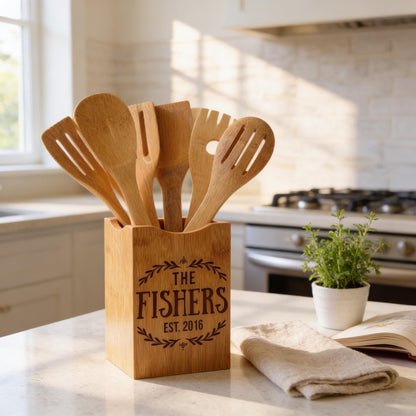 A wooden utensil holder on a kitchen counter, containing six wooden spoons, with a potted plant and a book nearby.