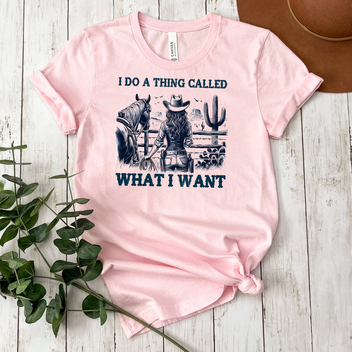 A pink t-shirt with a graphic design depicting a woman in cowboy attire, holding a horse, and the text "I DO A THING CALLED WHAT I WANT" printed on it.