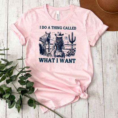 A pink t-shirt with a graphic design depicting a woman in cowboy attire, holding a horse, and the text "I DO A THING CALLED WHAT I WANT" printed on it.