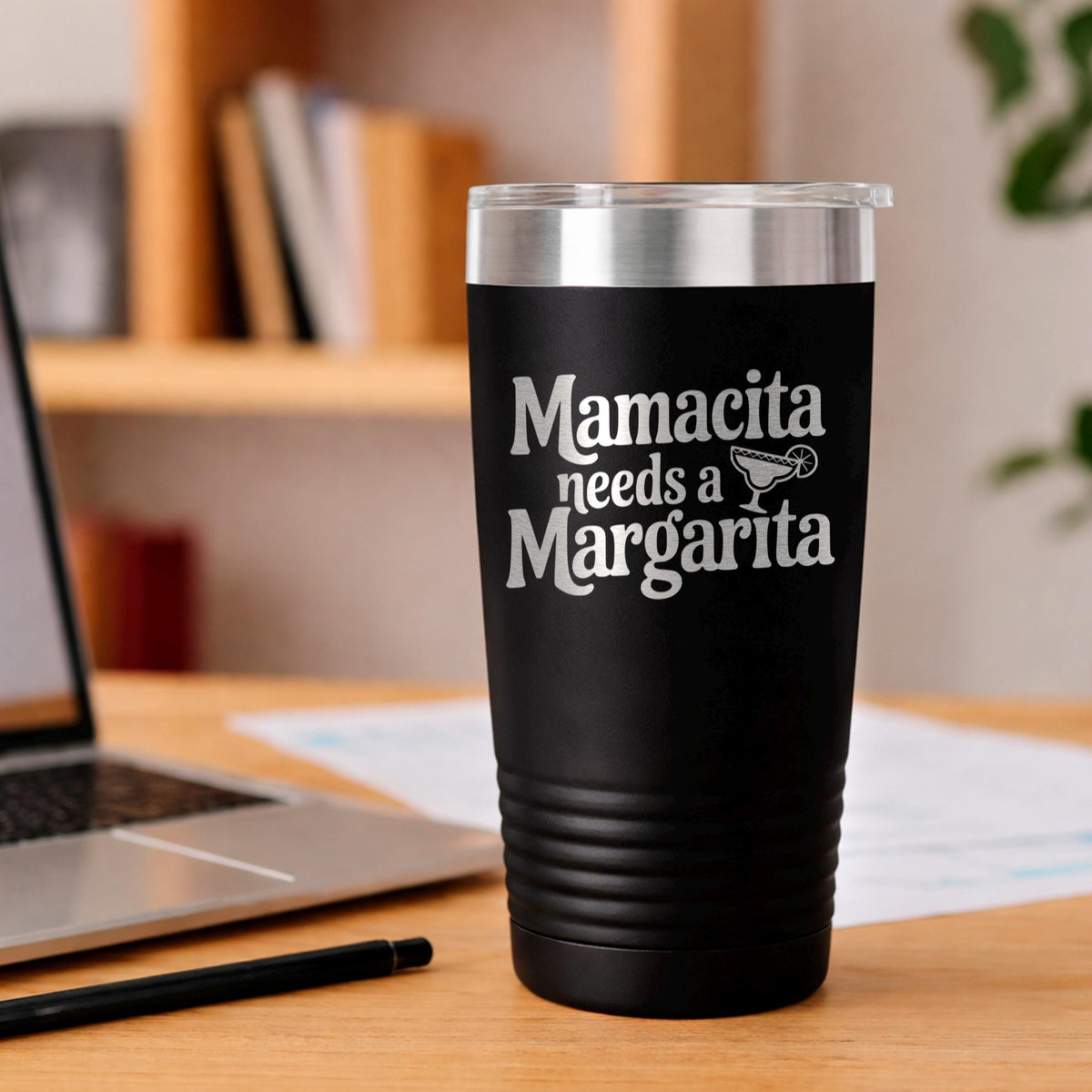 A black tumbler with the text "Mamacita needs a Margarita" printed on it, sitting on a wooden desk next to a laptop and a pen.