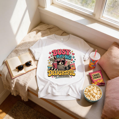 A white t-shirt with a colorful graphic design, a pair of sunglasses, a book, a bowl of popcorn, and a bag of chips are arranged on a window sill.