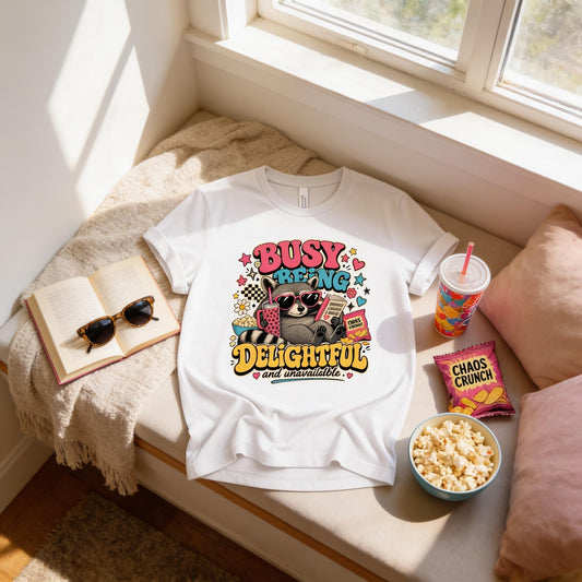 A white t-shirt with a colorful graphic design, a pair of sunglasses, a book, a bowl of popcorn, and a bag of chips are arranged on a window sill.