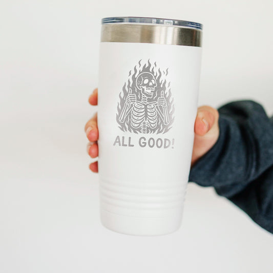A person's hand holding a white insulated tumbler with a printed design of a skeleton and flames, along with the text "ALL GOOD!"