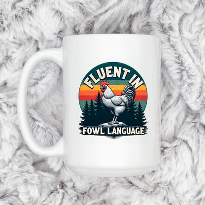 A white mug with a graphic design featuring a rooster and the text "FLUENT IN FOWL LANGUAGE" against a sunset background.